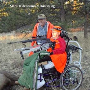Two hunters posing for photo with one hunter in wheelchair aiming down sights of